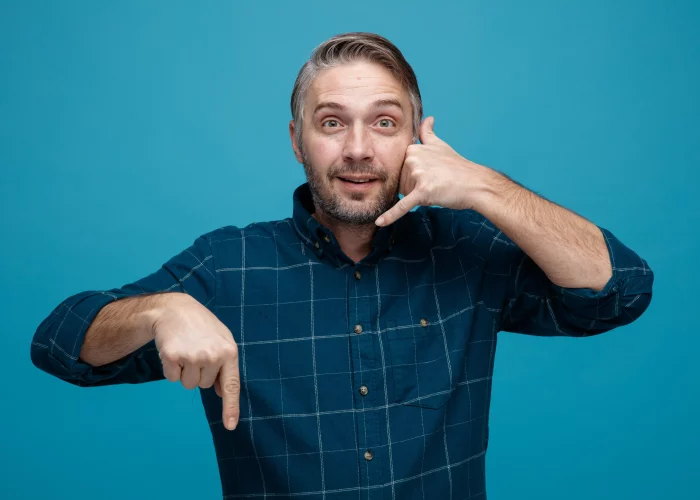 middle-age-man-with-grey-hair-dark-color-shirt-looking-camera-making-call-me-gesture-with-hand-pointing-with-index-finger-down-smiling-standing-blue-background